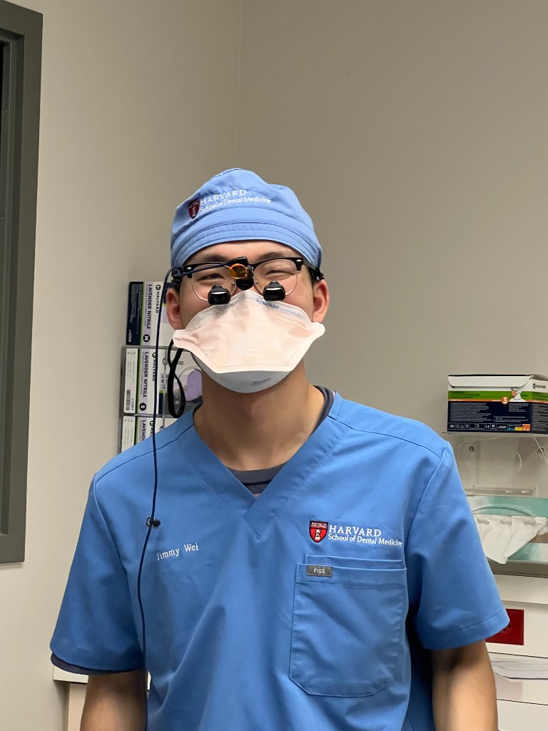 Dr. Jimmy Wei in surgical scrubs, cap, mask, and magnification loupes in a treatment room.